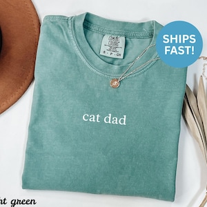 Könnte beinhalten: Hellgrünes T-Shirt mit dem weißen Text "cat dad". Das Shirt hat einen Rundhalsausschnitt und kurze Ärmel. Eine silberne Halskette mit Anhänger befindet sich auf dem Shirt. Das Shirt ist auf einer weißen Oberfläche gefaltet.