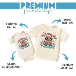 May include: Two cream-colored baby and toddler shirts. Each features a cartoon beaver wearing sunglasses in water, with the text "GOOD VIBES ONLY." The shirts are labeled "100% COTTON," "ULTRA COMFORTABLE," and "PREMIUM MATERIAL."