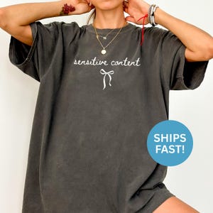 May include: Dark gray oversized t-shirt with the words "sensitive content" in a cursive font and a white bow graphic. The shirt has a vintage, worn look.