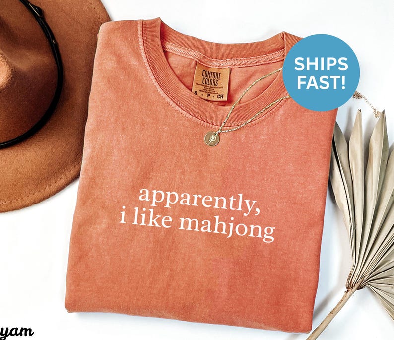 May include: A coral-colored t-shirt with the phrase "apparently, i like mahjong" printed in white. The shirt has a crew neck and a small tag on the collar. A gold necklace with a pendant is draped on the shirt.