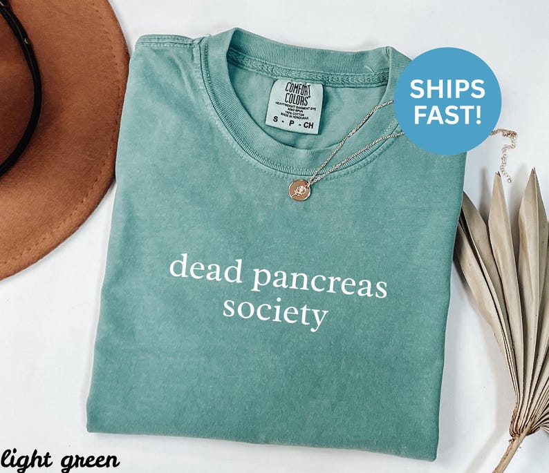 Puede incluir: Camiseta verde claro con el texto "dead pancreas society" en blanco. La camiseta tiene cuello redondo y mangas cortas. Un collar plateado con un colgante est&aacute; sobre la camiseta. La camiseta est&aacute; doblada sobre una superficie blanca.
