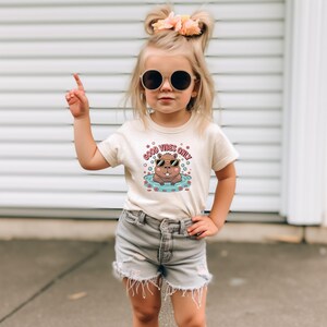 May include: A child wearing a cream-colored t-shirt with the text "GOOD VIBES ONLY" and a graphic of a cartoon beaver wearing sunglasses in water. The child is also wearing gray shorts and sunglasses.