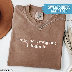 May include: Espresso-colored Comfort Colors t-shirt with the text "i may be wrong but i doubt it" in white. A silver necklace with a pendant rests on the shirt. The words "SWEATSHIRTS AVAILABLE" are in a blue banner.