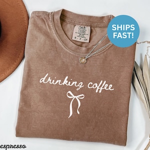 Peut inclure: T-shirt couleur expresso avec l'inscription "drinking coffee" en écriture blanche et un motif de nœud blanc. Le t-shirt a un col rond et des manches courtes. Un collier argenté avec un pendentif est drapé sur le t-shirt.