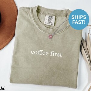 K&ouml;nnte beinhalten: Khakifarbenes T-Shirt mit dem Schriftzug "coffee first" in Wei&szlig;. Das Shirt hat einen Rundhalsausschnitt und ein kleines Etikett oben links. Eine silberne Halskette mit rosa Anh&auml;nger ist sichtbar. Ein brauner Hut ist links.
