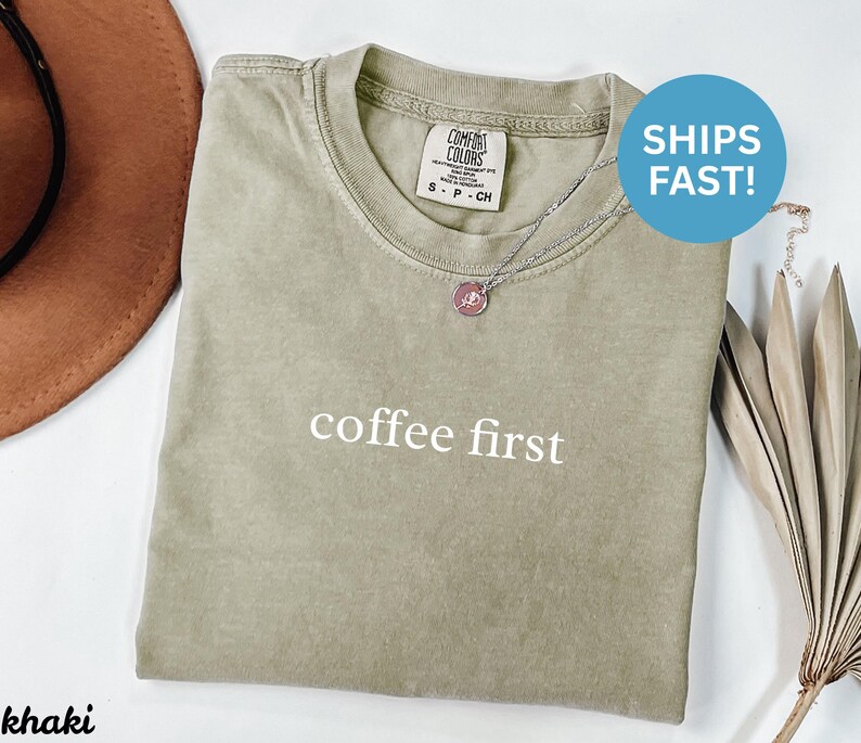 K&ouml;nnte beinhalten: Khakifarbenes T-Shirt mit dem Schriftzug "coffee first" in Wei&szlig;. Das Shirt hat einen Rundhalsausschnitt und ein kleines Etikett oben links. Eine silberne Halskette mit rosa Anh&auml;nger ist sichtbar. Ein brauner Hut ist links.