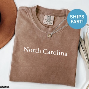 May include: Espresso-colored t-shirt with the text "North Carolina" in white. The shirt has a crew neck and a Comfort Colors tag. A silver necklace with a pendant is draped on the shirt.