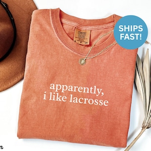 May include: A coral-colored t-shirt with the text "apparently, i like lacrosse" in white. The shirt has a crew neck and a small tag on the collar. A gold necklace with a pendant is visible. A brown hat is partially visible.