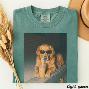 May include: A light green t-shirt with a graphic of a golden retriever wearing sunglasses and drinking an iced coffee. The t-shirt has a crew neck and short sleeves. The text "light green" is at the bottom right.