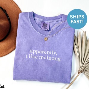 May include: A violet-colored t-shirt with the text "apparently, i like mahjong" in white. The shirt has a crew neck and is paired with a gold necklace. A brown hat and dried palm leaf are in the background.