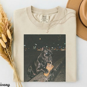 May include: Ivory Comfort Colors t-shirt with a graphic of a black dog wearing sunglasses and drinking from a coffee cup. The dog is sitting on a ledge with a city background. A silver necklace and a tan hat are also visible.