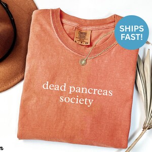 Puede incluir: Una camiseta de color coral con las palabras "dead pancreas society" en blanco. La camiseta tiene cuello redondo y una peque&ntilde;a etiqueta en el cuello. Se ve un collar dorado con un colgante. Un sombrero marr&oacute;n est&aacute; en el fondo.