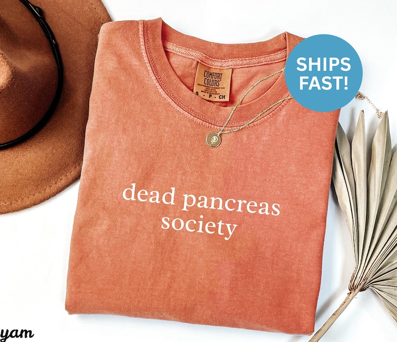 Puede incluir: Una camiseta de color coral con las palabras "dead pancreas society" en blanco. La camiseta tiene cuello redondo y una peque&ntilde;a etiqueta en el cuello. Se ve un collar dorado con un colgante. Un sombrero marr&oacute;n est&aacute; en el fondo.