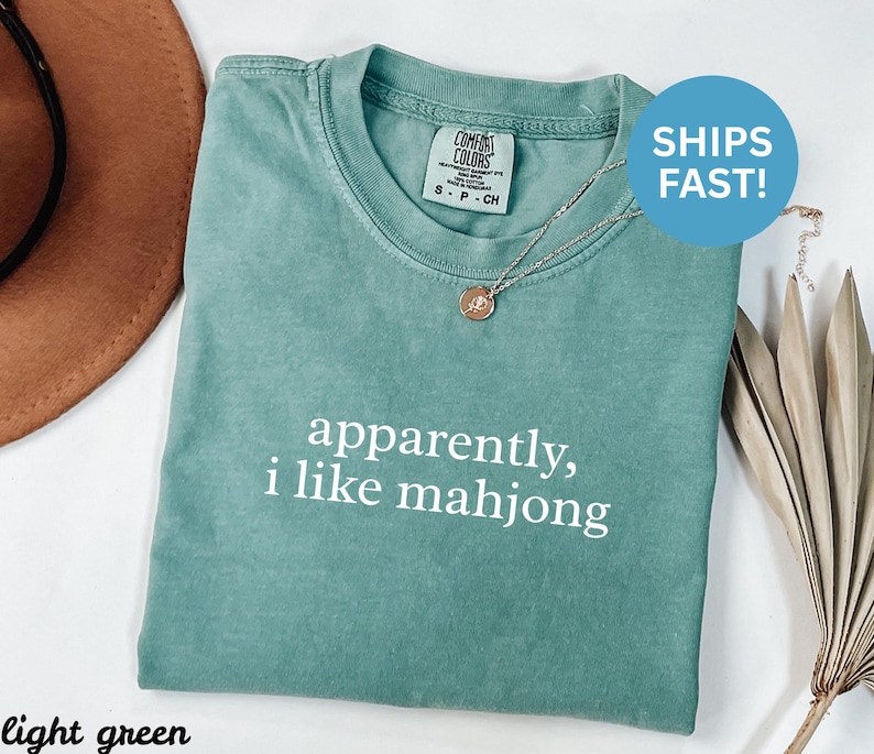 May include: Light green t-shirt with the text "apparently, I like mahjong" in white. The shirt has a crew neck and is made of a soft material. A silver necklace with a pendant is visible.
