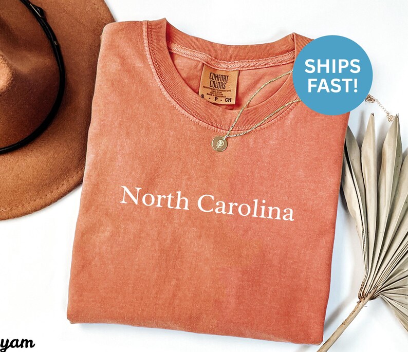 May include: A coral-colored t-shirt with the words "North Carolina" printed in white. The shirt has a crew neck and a small tag on the collar. A gold necklace with a pendant is draped on the shirt. A brown hat is visible in the background.