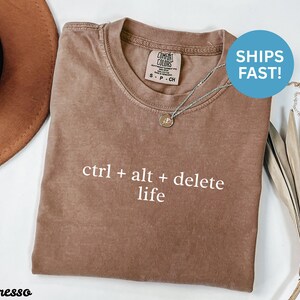 以下が含まれることがあります： 「ctrl + alt + delete life」の白い文字が入ったエスプレッソカラーのTシャツ。Tシャツはクルーネックと半袖です。小さなペンダントが付いたシルバーのネックレスがTシャツの上にあります。