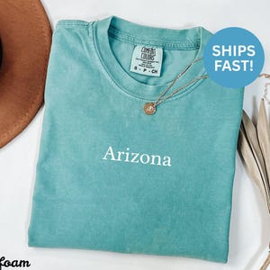 May include: Seafoam green t-shirt with the word "Arizona" printed in white. The shirt has a crew neck and short sleeves. A gold necklace with a pendant is on the shirt. A brown hat is in the background.