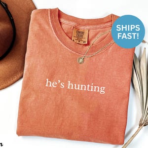 May include: A coral-colored t-shirt with the phrase "he's hunting" printed in white. The shirt has a crew neck and a small tag on the collar. A gold necklace with a pendant is visible. A brown hat is in the background.