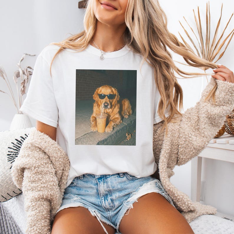 Woman wearing white t-shirt with graphic of golden retriever in sunglasses holding coffee.