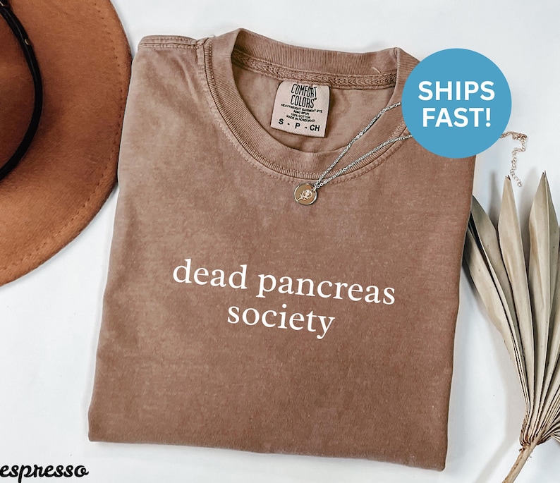 Puede incluir: Camiseta marr&oacute;n con la frase "dead pancreas society" en blanco. La camiseta tiene cuello redondo y mangas cortas. Un collar plateado con un peque&ntilde;o colgante est&aacute; sobre la camiseta. Un sombrero marr&oacute;n es visible en el fondo.
