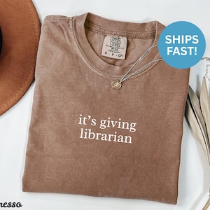 Op de afbeelding: Bruin T-shirt met de witte tekst "it's giving librarian". Het shirt heeft een ronde hals en korte mouwen. Een zilveren ketting met een kleine hanger is zichtbaar. Het shirt is gelabeld met "Comfort Colors".