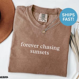 Może przedstawiać: Koszulka w kolorze espresso z białym napisem "forever chasing sunsets". Koszulka ma okrągły dekolt i krótkie rękawy. Srebrny naszyjnik z małą zawieszką leży na koszulce. Brązowy kapelusz jest w tle.