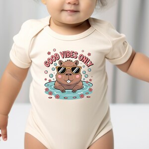 May include: A beige baby bodysuit with a graphic of a cartoon beaver wearing sunglasses in a pool. The text "GOOD VIBES ONLY" is above the beaver. Pink bubbles and flowers surround the beaver.