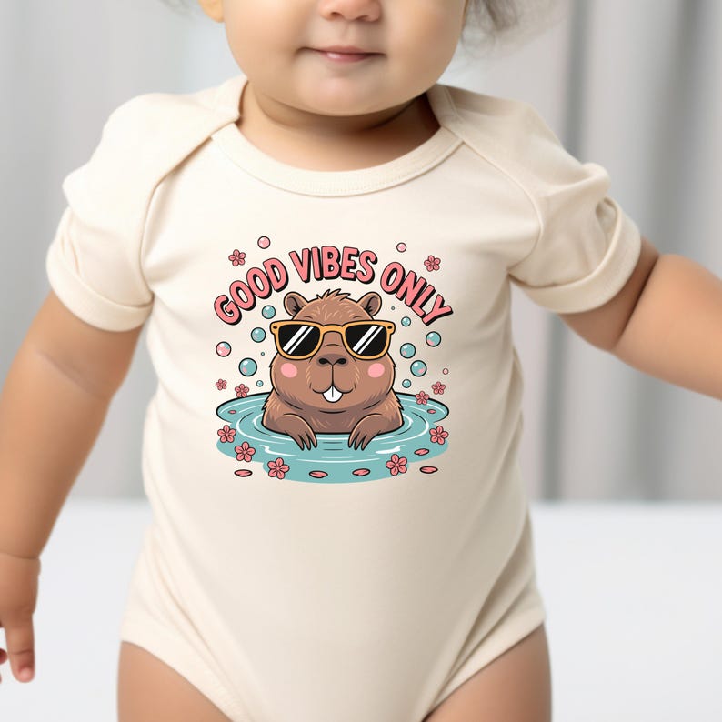 May include: A beige baby bodysuit with a graphic of a cartoon beaver wearing sunglasses in a pool. The text "GOOD VIBES ONLY" is above the beaver. Pink bubbles and flowers surround the beaver.