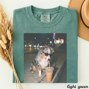 Puede incluir: Camiseta Comfort Colors verde claro con un gráfico de un perro con gafas de sol bebiendo café helado con una pajita. El perro está sentado en una acera por la noche. Un sombrero marrón es visible en el fondo.