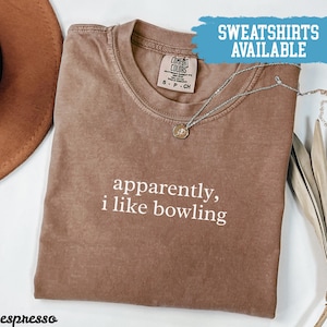 May include: Espresso-coloured t-shirt with the phrase "apparently, i like bowling" in white text. A silver necklace with a small pendant rests on the shirt. The shirt is labelled "Comfort Colors" and "Sweatshirts Available" is written in a blue banner.