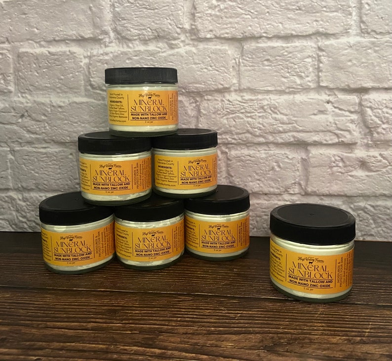 May include: A collection of seven small jars of mineral sunscreen with black lids. Each jar has a label with the words "Mineral Sunblock" and "Made with Tallow and Non-Nano Zinc Oxide". The jars are arranged on a wooden surface.