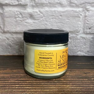 May include: A small glass jar with a black lid, containing a creamy, off-white substance. A yellow label lists ingredients like organic olive oil and beeswax. The text "MINERAL SUNBLOOM" is visible on the label.