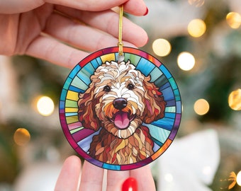 May include: A round, stained-glass style ornament featuring a smiling dog with brown and cream fur. The ornament has a colorful border and a gold string for hanging. The dog's tongue is visible.