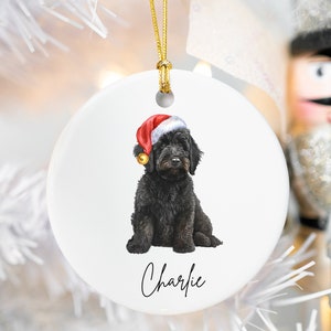 Könnte beinhalten: Ein weißer Keramik-Weihnachtsbaumschmuck mit einem schwarzen Hund, der einen roten Weihnachtsmann-Hut trägt. Der Name des Hundes, "Charlie", ist unten auf dem Ornament in Kursivschrift geschrieben.