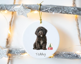 May include: A white ceramic Christmas ornament featuring a watercolor illustration of a black dog with the name "Teddy" written below. A small snow globe with a Christmas tree is next to the dog. The ornament hangs from a gold string.