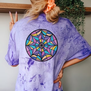 May include: Purple tie-dye t-shirt with a colorful mandala design on the back. The mandala features a symmetrical pattern with shades of blue, yellow, and purple. The shirt has a relaxed fit and short sleeves.