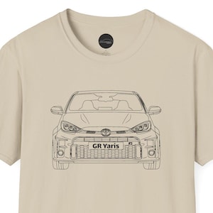 Puede incluir: Camiseta beige con un dibujo lineal negro de un coche GR Yaris. La vista frontal del coche está centrada en la camiseta, con el texto "GR Yaris" visible en la parrilla. La camiseta tiene cuello redondo y mangas cortas.