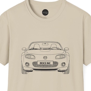 Può includere: T-shirt beige con un disegno a tratto nero di un'auto. L'auto ha i fari, una griglia e il testo "MX5 NC" sul davanti. La maglietta è di colore neutro e ha uno scollo tondo.