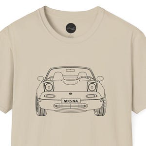 May include: A beige t-shirt with a black outline of a convertible car. The car design includes headlights, wheels, and the text "MKS NA". The shirt's neutral tone makes it versatile for different outfits and occasions.