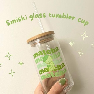 May include: A clear glass tumbler with a bamboo lid and a green and white illustration of a cartoon character holding a cup of matcha. The tumbler has the word "matcha" repeated multiple times on the side.