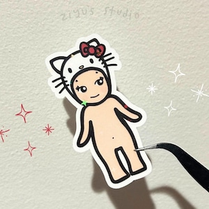 May include: A sticker featuring a cartoon figure wearing a white Hello Kitty-themed hat with a red bow. The figure has a light skin tone and is outlined in black. The sticker is being held by tweezers.