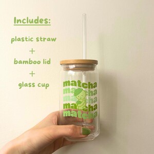 May include: A glass cup with a bamboo lid and a green plastic straw. The cup has a green and white graphic of a person holding a matcha cup with the word "matcha" repeated on the cup.