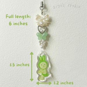 K&ouml;nnte beinhalten: Ein 15 cm langer Anh&auml;nger mit einem Hasen-Design. Der Anh&auml;nger hat eine cremefarbene Schleife, eine herzf&ouml;rmige Perle, einen gr&uuml;nen Schmetterling, eine wei&szlig;e Blume und einen transparenten Acryl-Anh&auml;nger in Hasenform. Der Hasenanh&auml;nger ist 3,8 cm hoch und 3 cm breit.