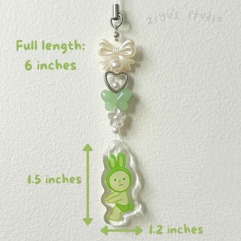 K&ouml;nnte beinhalten: Ein 15 cm langer Anh&auml;nger mit einem Hasen-Design. Der Anh&auml;nger hat eine cremefarbene Schleife, eine herzf&ouml;rmige Perle, einen gr&uuml;nen Schmetterling, eine wei&szlig;e Blume und einen transparenten Acryl-Anh&auml;nger in Hasenform. Der Hasenanh&auml;nger ist 3,8 cm hoch und 3 cm breit.