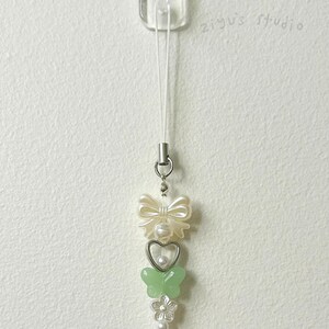 May include: A white phone charm with a white bow, a heart with a pearl, a green butterfly, and a pearl flower. The charm is attached to a white string.