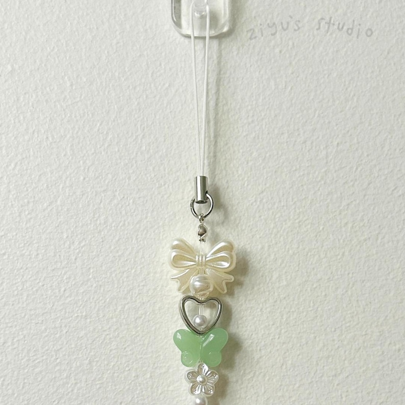 May include: A white phone charm with a white bow, a heart with a pearl, a green butterfly, and a pearl flower. The charm is attached to a white string.