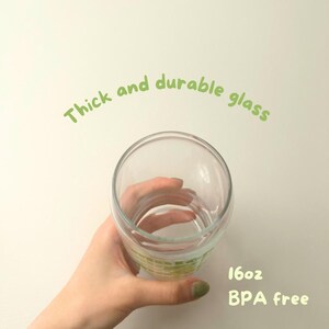 May include: A clear glass cup with a thick base, held in a hand. The cup is 16 oz and BPA free. The text "Thick and durable glass" is written in green above the cup.