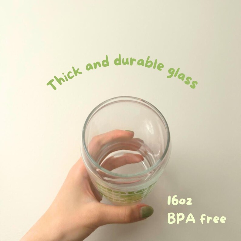 May include: A clear glass cup with a thick base, held in a hand. The cup is 16 oz and BPA free. The text "Thick and durable glass" is written in green above the cup.