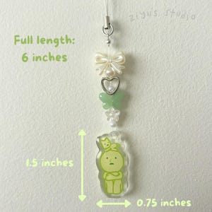 May include: A green and white charm with a cartoon character and a cat on its head. The charm is attached to a white string with a bow, butterflies, and flowers. The charm is 1.5 inches tall and 0.75 inches wide. Full length: 6 inches.