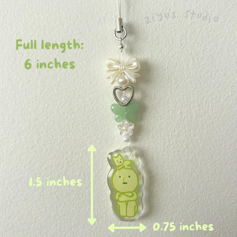 May include: A green and white charm with a cartoon character and a cat on its head. The charm is attached to a white string with a bow, butterflies, and flowers. The charm is 1.5 inches tall and 0.75 inches wide. Full length: 6 inches.